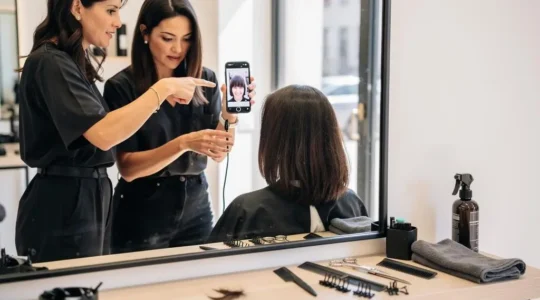 Une coiffeuse montre une photo de référence à sa cliente assise devant le miroir d'un salon de coiffure lumineux.