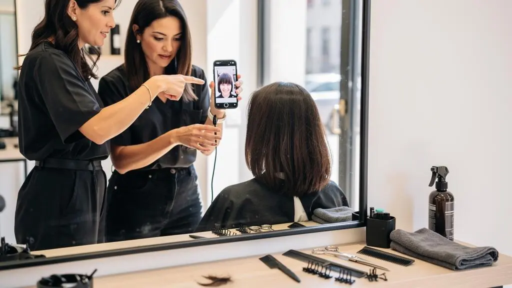 Une coiffeuse montre une photo de référence à sa cliente assise devant le miroir d'un salon de coiffure lumineux.