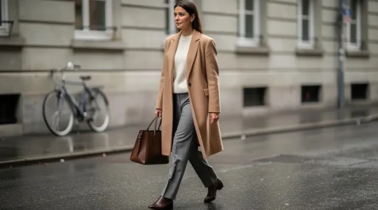 Une femme marche dans une rue suisse au style épuré, vêtue d'un manteau en laine camel, d'un pull crème et d'un pantalon gris ajusté, sous une lumière douce.