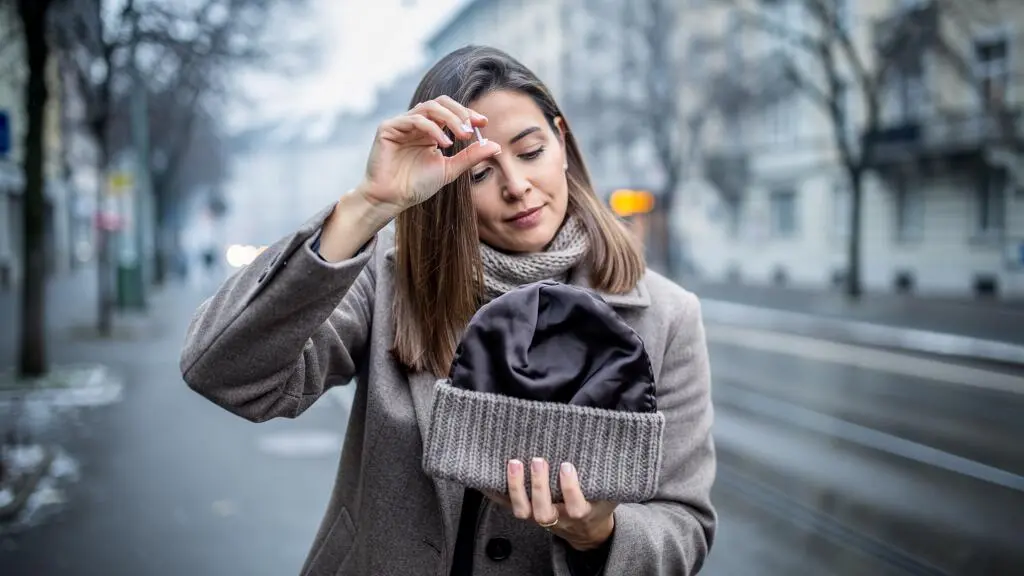 Une femme applique un soin protecteur sur ses longueurs avant de mettre son bonnet doublé sur un trottoir suisse en hiver.