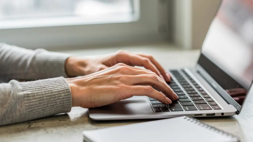 Mains aux ongles courts avec une manucure nude rosé nette tapant sur un clavier dans un bureau lumineux